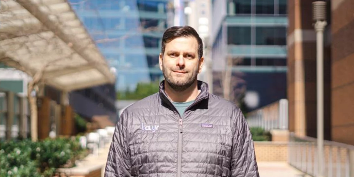 Man outdoors wearing a Layr jacket, looking into the camera; modern office buildings in the background.